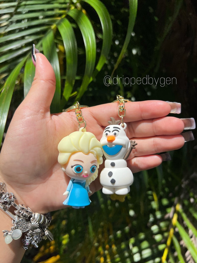 Op de afbeelding: Een set van twee Frozen-sleutelhangers met Elsa en Olaf. Elsa draagt een blauwe jurk en heeft blond haar. Olaf is een sneeuwpop met een wortelneus en een blauwe sjaal. De sleutelhangers zijn bevestigd aan gouden kettingen.