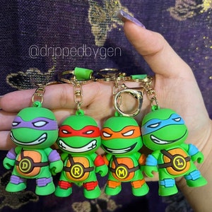 Op de afbeelding: Vier sleutelhangers van de Teenage Mutant Ninja Turtles. Elke sleutelhanger heeft een andere schildpad: Donatello, Raphael, Michelangelo en Leonardo. Elke schildpad is groen met een anders gekleurd masker en bandana. De sleutelhangers zijn gemaakt van plastic en hebben een gouden metalen ring.