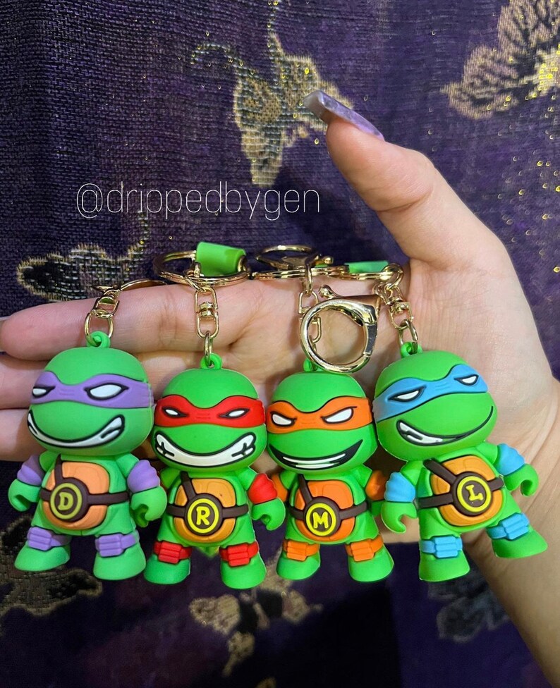 Op de afbeelding: Vier sleutelhangers van de Teenage Mutant Ninja Turtles. Elke sleutelhanger heeft een andere schildpad: Donatello, Raphael, Michelangelo en Leonardo. Elke schildpad is groen met een anders gekleurd masker en bandana. De sleutelhangers zijn gemaakt van plastic en hebben een gouden metalen ring.