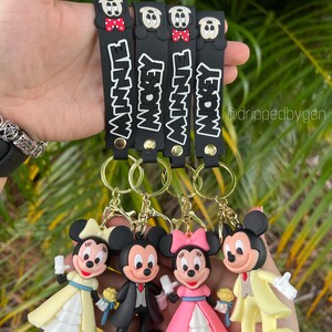 Op de afbeelding: Vier zwarte sleutelhangers met Mickey Mouse en Minnie Mouse figuren. Elke sleutelhanger heeft een zwarte band met witte tekst die "Mickey" of "Minnie" zegt. De figuren zijn gekleed in trouwjurken.