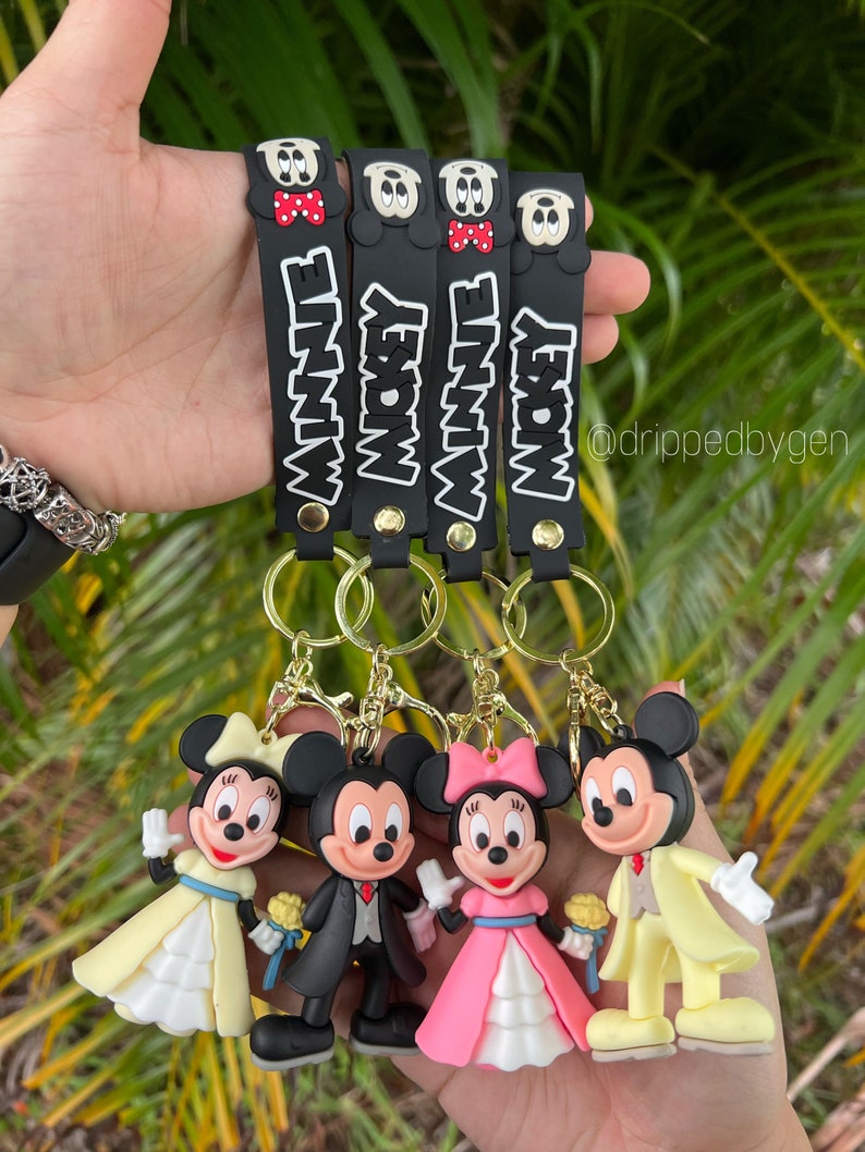 Op de afbeelding: Vier zwarte sleutelhangers met Mickey Mouse en Minnie Mouse figuren. Elke sleutelhanger heeft een zwarte band met witte tekst die "Mickey" of "Minnie" zegt. De figuren zijn gekleed in trouwjurken.