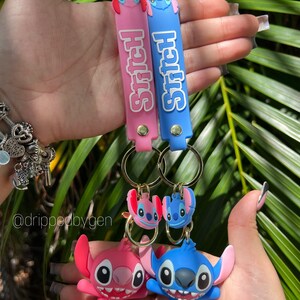 Op de afbeelding: Twee Stitch-sleutelhangers met blauwe en roze siliconen banden. De banden hebben het woord "Stitch" erop gedrukt. De sleutelhangers hebben 3D Stitch-figuren met blauwe en roze accenten.