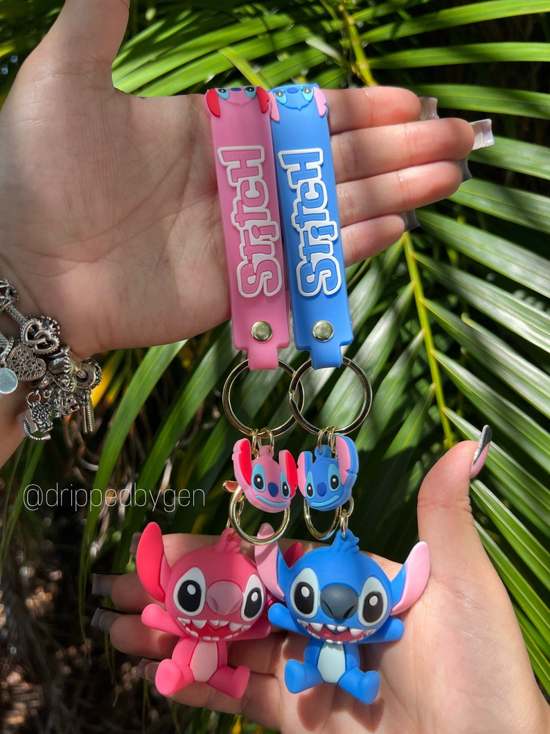Op de afbeelding: Twee Stitch-sleutelhangers met blauwe en roze siliconen banden. De banden hebben het woord "Stitch" erop gedrukt. De sleutelhangers hebben 3D Stitch-figuren met blauwe en roze accenten.