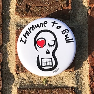 May include: White button with a black and white skull illustration with a red heart for an eye. The text around the skull reads "Immune to Bull".