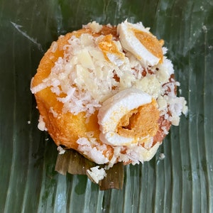 May include: A round, golden-brown pastry topped with shredded white coconut and sliced, hard-boiled eggs with orange yolks. The pastry rests on a large, green banana leaf, creating a natural presentation.