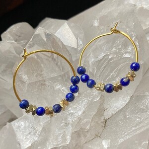 May include: Gold hoop earrings with blue lapis lazuli beads and gold spacers. The earrings are displayed on a white crystal cluster.