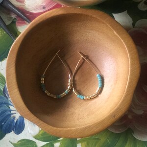 May include: A pair of gold-colored teardrop earrings with turquoise and gold beads. The earrings are displayed in a wooden bowl. The beads are evenly spaced along the bottom curve of the earrings, creating a delicate and stylish accessory.