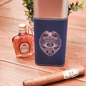 May include: A blue leather cigar case with a silver top and a police officer badge design. The badge has the text "Police Officer", "Gwinnett County", "Police", and "314". The case is next to a small bottle of Crown Royal Vanilla flavored whiskey and a brown cigar.