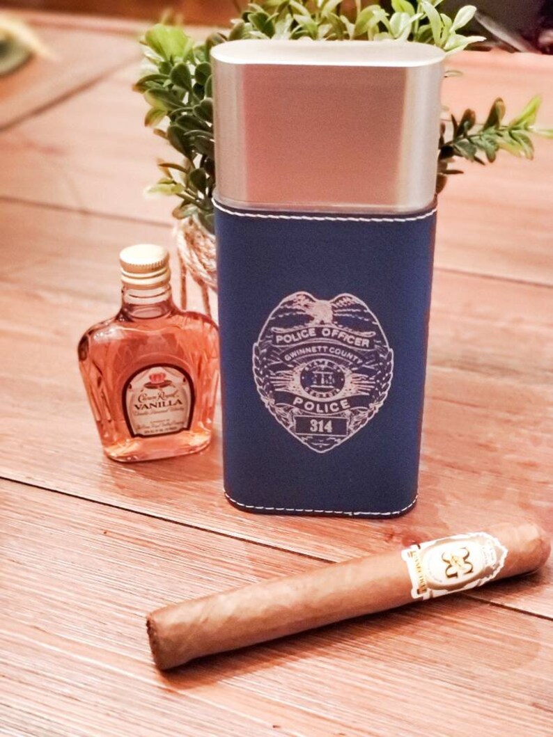 May include: A blue leather cigar case with a silver top and a police officer badge design. The badge has the text "Police Officer", "Gwinnett County", "Police", and "314". The case is next to a small bottle of Crown Royal Vanilla flavored whiskey and a brown cigar.