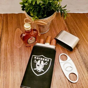 May include: A black leather cigar case with the Las Vegas Raiders logo and a silver metal cigar cutter. The case has a silver metal trim and is sitting on a wooden surface.