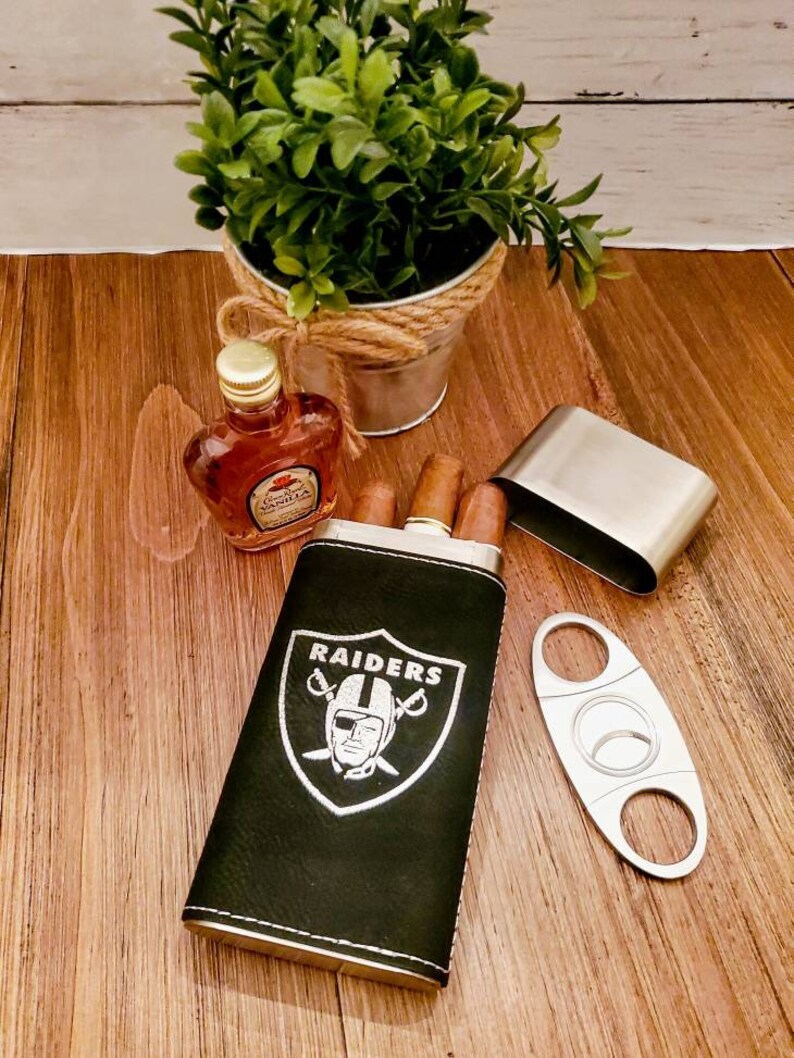 May include: A black leather cigar case with the Las Vegas Raiders logo and a silver metal cigar cutter. The case has a silver metal trim and is sitting on a wooden surface.