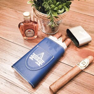 May include: A blue leather cigar case with the word "AMIZIAH" engraved on it. The case is open and contains two cigars. A small bottle of Canadian Club Vanilla liqueur is also visible in the image.