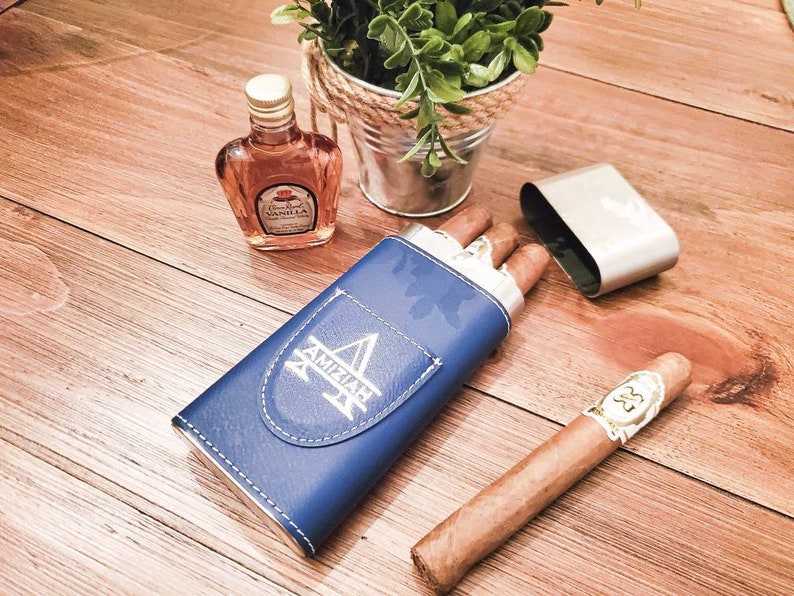 May include: A blue leather cigar case with the word "AMIZIAH" engraved on it. The case is open and contains two cigars. A small bottle of Canadian Club Vanilla liqueur is also visible in the image.