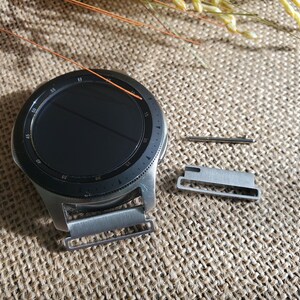 Pu&ograve; includere: Un orologio intelligente argentato con un quadrante nero e un cinturino in metallo argentato. L'orologio &egrave; appoggiato su una superficie di juta marrone con un piccolo perno in metallo argentato e un connettore per cinturino in metallo argentato.