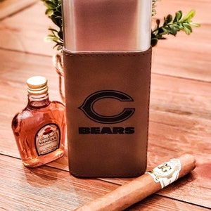 May include: A brown leather cigar case with a silver metal insert and the Chicago Bears logo. The case is sitting on a wooden surface with a bottle of Crown Royal Vanilla and a cigar.