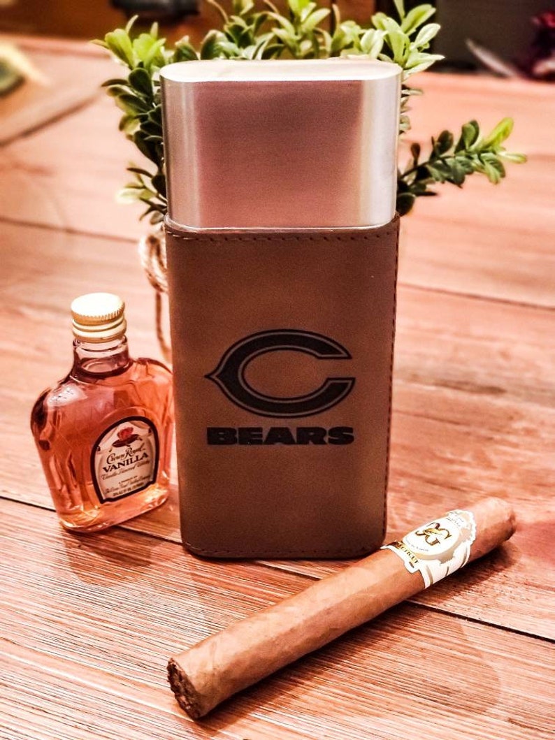May include: A brown leather cigar case with a silver metal insert and the Chicago Bears logo. The case is sitting on a wooden surface with a bottle of Crown Royal Vanilla and a cigar.