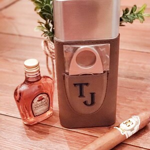 May include: A brown leather cigar case with a silver top, a cigar cutter, and the initials "TJ" embossed on the front. A small bottle of Crown Royal Vanilla liqueur and a brown cigar are also pictured.