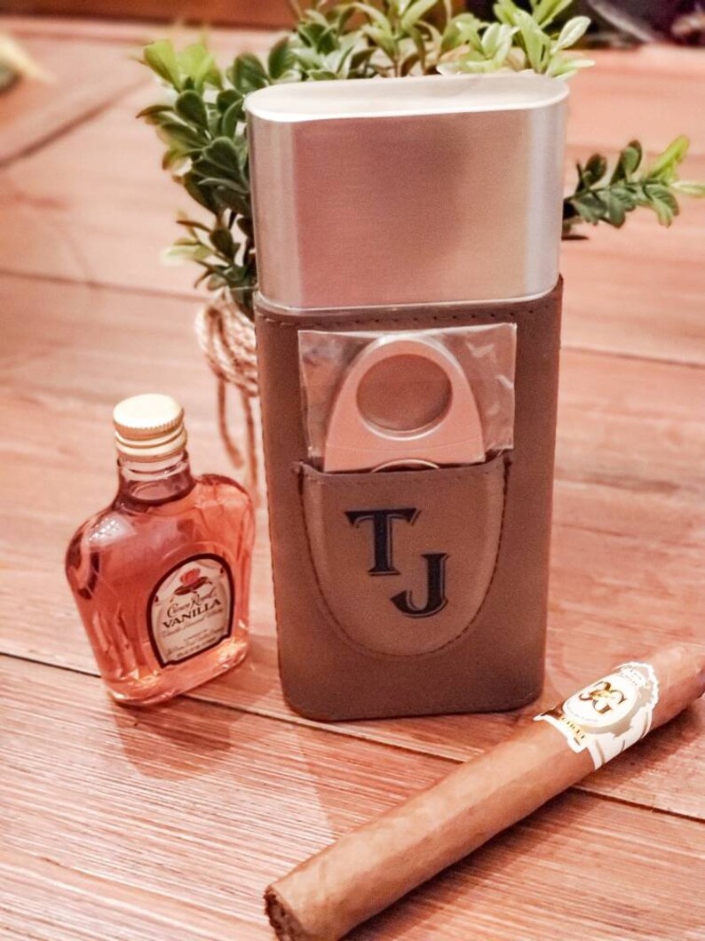 May include: A brown leather cigar case with a silver top, a cigar cutter, and the initials "TJ" embossed on the front. A small bottle of Crown Royal Vanilla liqueur and a brown cigar are also pictured.