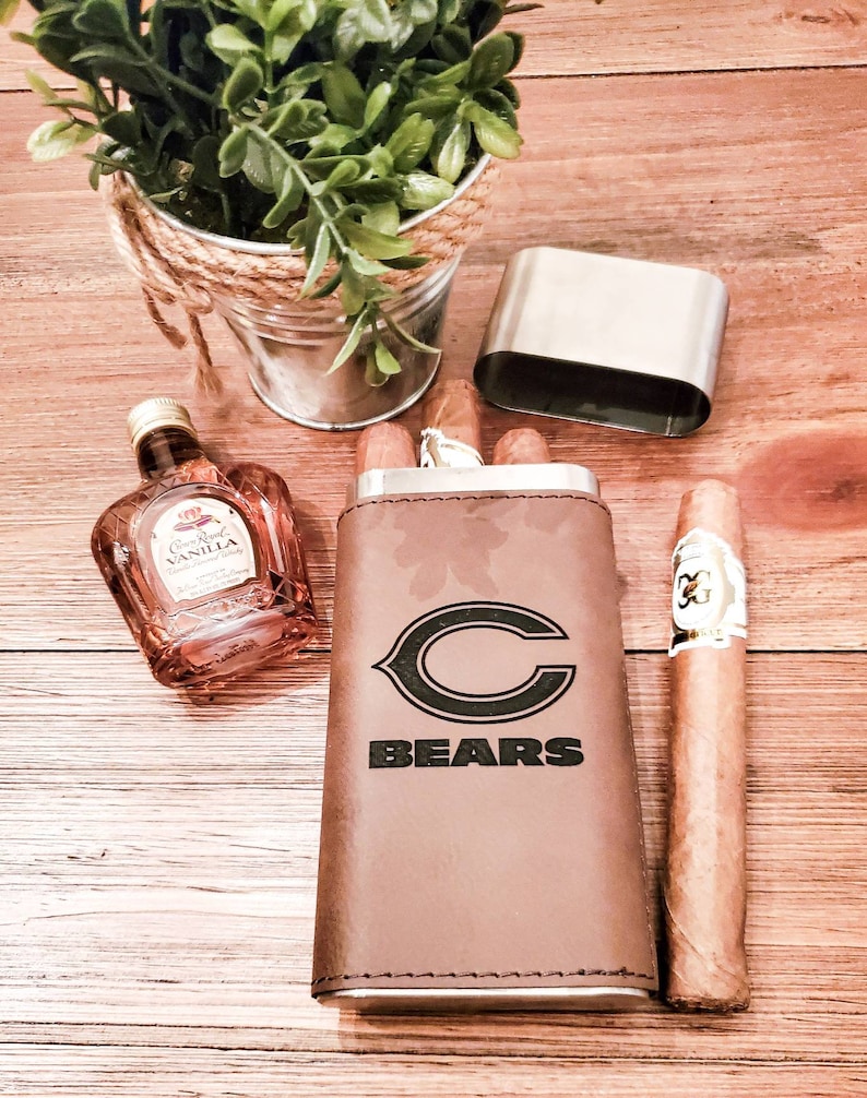 May include: A brown leather cigar case with a metal base and a Chicago Bears logo. The case is open and has three cigars inside. A small bottle of Crown Royal Vanilla whiskey is also in the image.