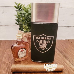 May include: A black leather cigar case with a silver metal interior and the Raiders logo embroidered on the front. The case is sitting on a wooden surface next to a small bottle of liquor and a cigar.