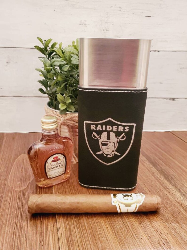 May include: A black leather cigar case with a silver metal interior and the Raiders logo embroidered on the front. The case is sitting on a wooden surface next to a small bottle of liquor and a cigar.