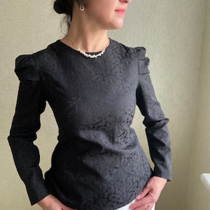 May include: A black long-sleeved blouse with a floral pattern and a fitted waist. The blouse has puffed sleeves and a round neckline. The model is wearing a pearl necklace.
