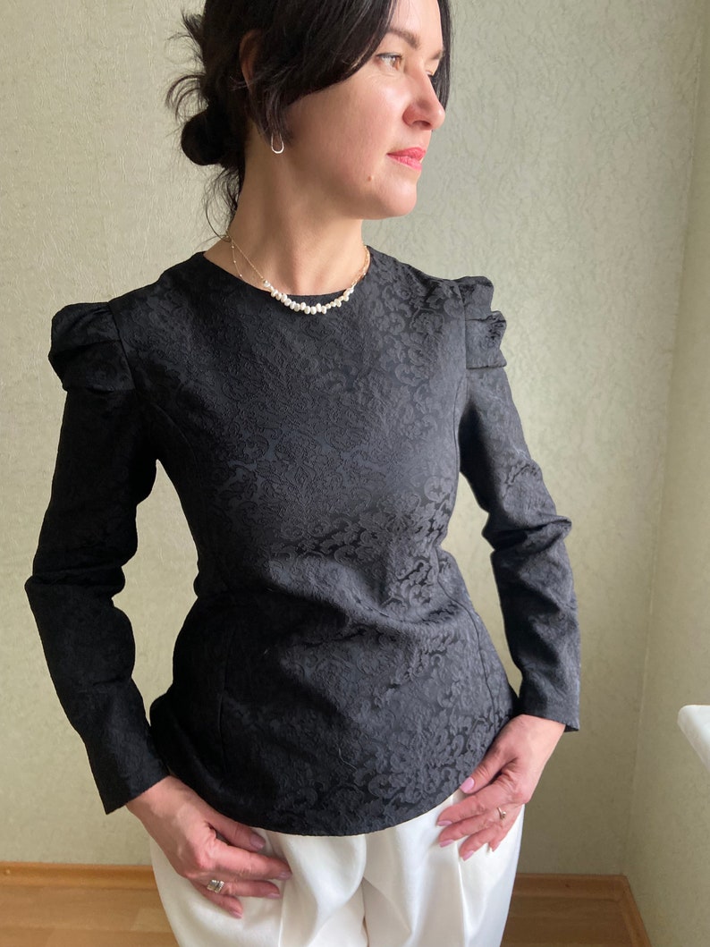May include: A black long-sleeved blouse with a floral pattern and a fitted waist. The blouse has puffed sleeves and a round neckline. The model is wearing a pearl necklace.