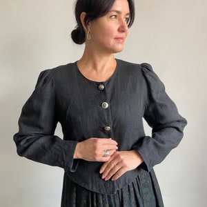 Puede incluir: Una chaqueta negra de lino con cuello redondo y cierre de botones. La chaqueta tiene mangas abullonadas y una cintura ajustada. La modelo lleva la chaqueta con una falda floral negra y verde.
