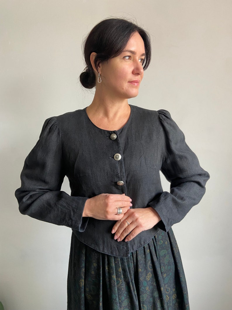 Puede incluir: Una chaqueta negra de lino con cuello redondo y cierre de botones. La chaqueta tiene mangas abullonadas y una cintura ajustada. La modelo lleva la chaqueta con una falda floral negra y verde.