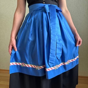Puede incluir: Una falda de dirndl azul con un ribete a rayas blanco y rojo y bordados florales. La falda tiene una cinta negra atada a la cintura y una falda interior negra.