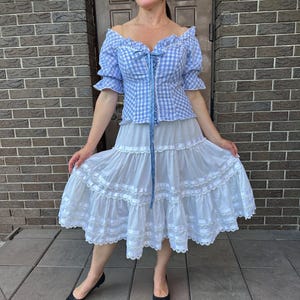 Puede incluir: Una blusa de hombros descubiertos con estampado de cuadros vichy en azul claro y blanco, mangas cortas abullonadas y cordones frontales con cinta azul, junto a una falda blanca midi con varios niveles horizontales, encaje y cintas.