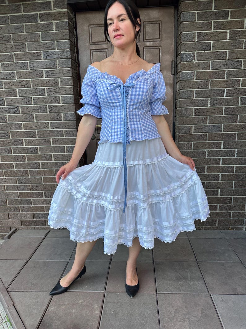 Puede incluir: Una blusa de hombros descubiertos con estampado de cuadros vichy en azul claro y blanco, mangas cortas abullonadas y cordones frontales con cinta azul, junto a una falda blanca midi con varios niveles horizontales, encaje y cintas.