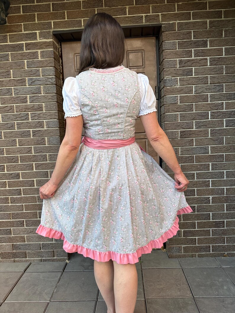 May include: A grey dirndl dress featuring a small pink floral pattern, a white blouse with short puffed sleeves, a pink waist sash, and a pink ruffled hem. The bodice has a fitted design with a lace trim along the neckline, and the skirt is pleated.