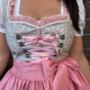 May include: A dirndl dress featuring a grey bodice with a pink floral pattern, a square neckline trimmed with white and pink lace, and a pink ribbon lace-up front with decorative metal eyelets. The dress includes a solid pink apron with a large bow.
