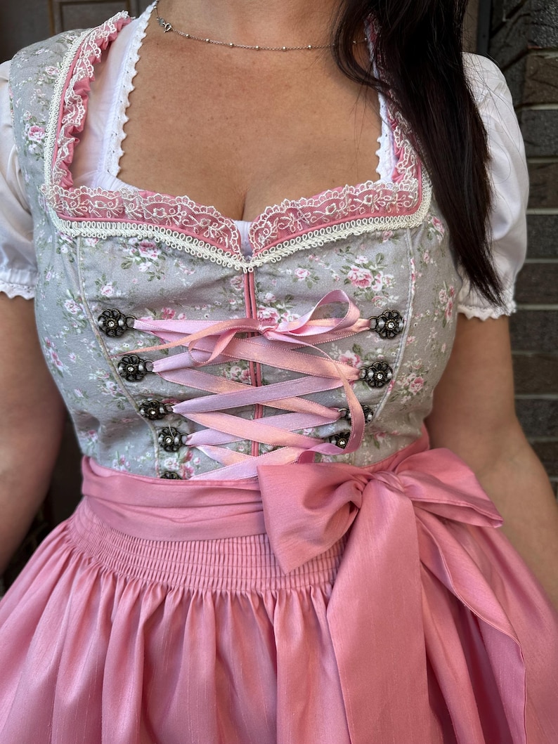 May include: A dirndl dress featuring a grey bodice with a pink floral pattern, a square neckline trimmed with white and pink lace, and a pink ribbon lace-up front with decorative metal eyelets. The dress includes a solid pink apron with a large bow.