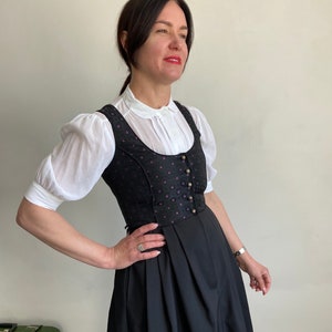 Puede incluir: Un vestido dirndl negro con un corpi&ntilde;o que presenta un sutil estampado en tonos morado y negro, escote redondo y una hilera de botones met&aacute;licos. El vestido se lleva sobre una blusa blanca de mangas cortas abullonadas y cuello redondo.