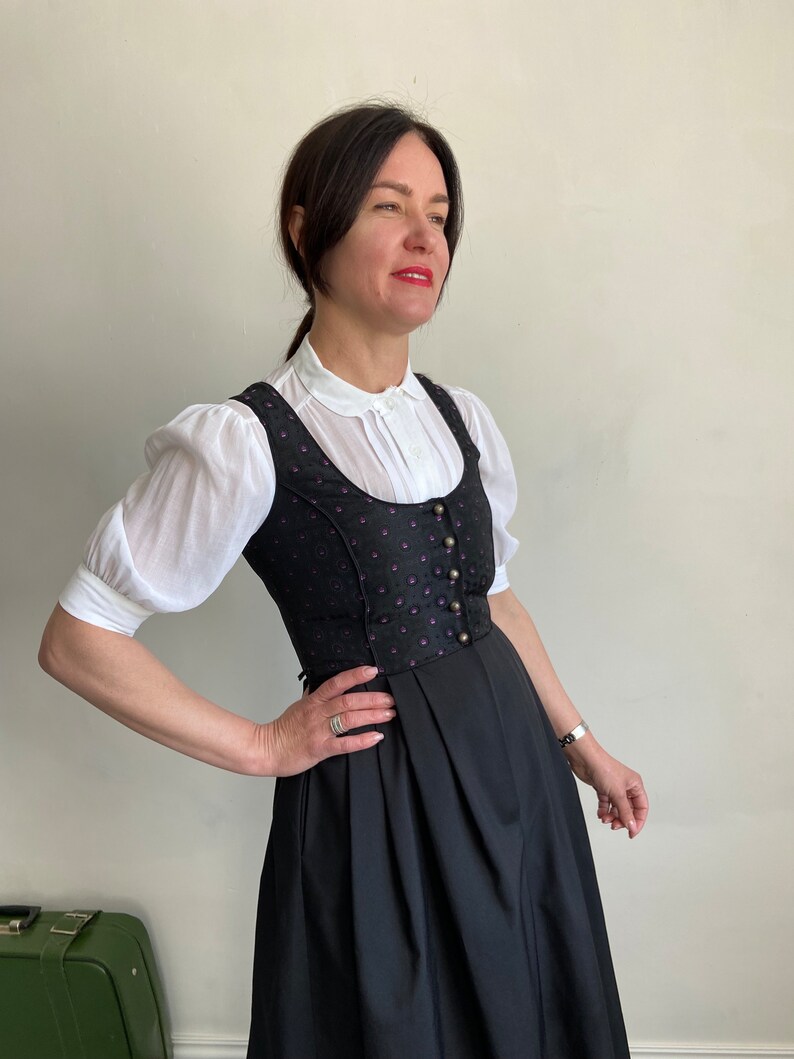 Puede incluir: Un vestido dirndl negro con un corpi&ntilde;o que presenta un sutil estampado en tonos morado y negro, escote redondo y una hilera de botones met&aacute;licos. El vestido se lleva sobre una blusa blanca de mangas cortas abullonadas y cuello redondo.