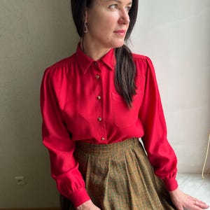 Puede incluir: Una mujer con una camisa roja de manga larga con cuello abotonado y una falda de cuadros marrones y verdes.