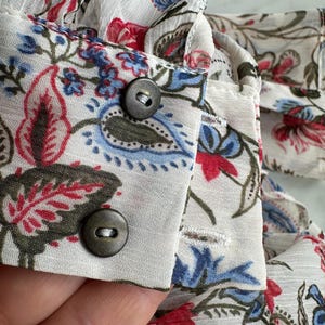 May include: Close-up of a white fabric with a floral print. The fabric has two brown buttons and a white ruffle along the top edge.