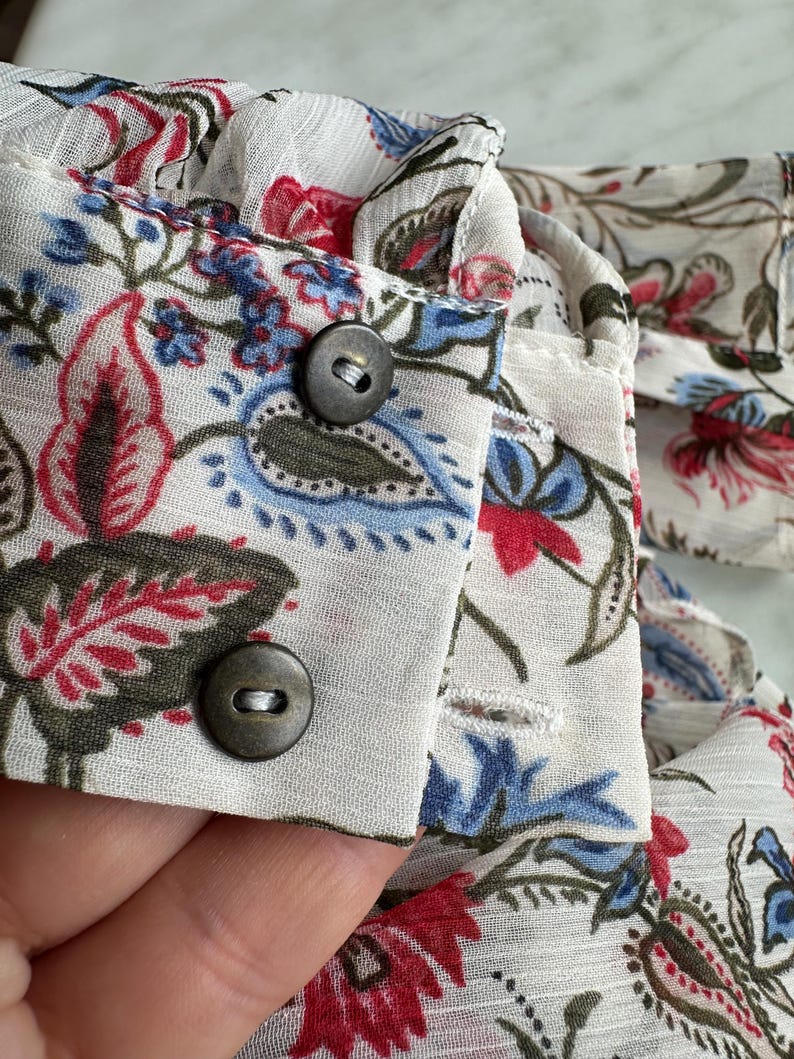May include: Close-up of a white fabric with a floral print. The fabric has two brown buttons and a white ruffle along the top edge.