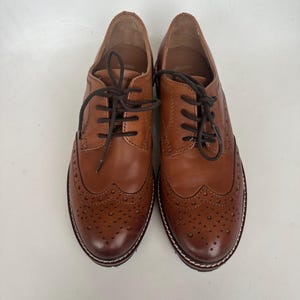 May include: A pair of brown leather brogue shoes with dark brown laces. The shoes have decorative perforations on the toe and sides, and a white stitched sole. The shoes are displayed on a white background.