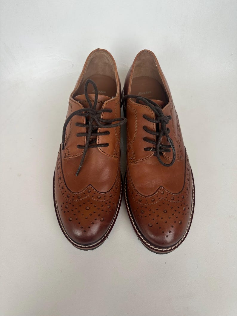 May include: A pair of brown leather brogue shoes with dark brown laces. The shoes have decorative perforations on the toe and sides, and a white stitched sole. The shoes are displayed on a white background.