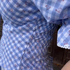 Puede incluir: Una blusa de cuadros vichy en azul claro y blanco con un peque&ntilde;o estampado floral rojo. La prenda incluye un panel lateral con detalles de nido de abeja fruncido y mangas cortas terminadas con un delicado borde de encaje festoneado blanco.