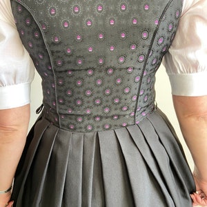 Puede incluir: Un vestido dirndl con un corpi&ntilde;o gris oscuro con un patr&oacute;n floral repetido en negro y magenta, combinado con una falda plisada gris oscuro lisa y una blusa blanca de manga corta. El corpi&ntilde;o tiene un ajuste estructurado con costuras visibles.