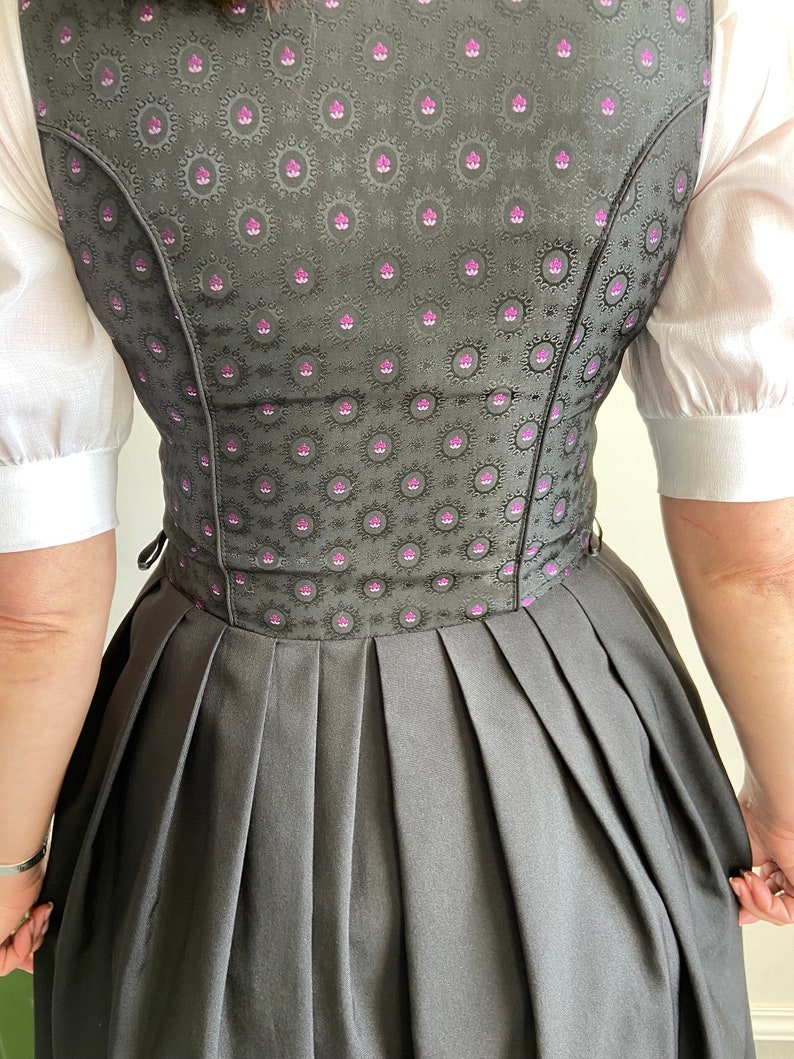 Puede incluir: Un vestido dirndl con un corpi&ntilde;o gris oscuro con un patr&oacute;n floral repetido en negro y magenta, combinado con una falda plisada gris oscuro lisa y una blusa blanca de manga corta. El corpi&ntilde;o tiene un ajuste estructurado con costuras visibles.