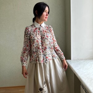 May include: A woman wearing a white collared blouse with a floral print and a beige skirt with embroidery. The blouse has long sleeves and a button-up front. The skirt is a-line and falls to the woman's ankles.