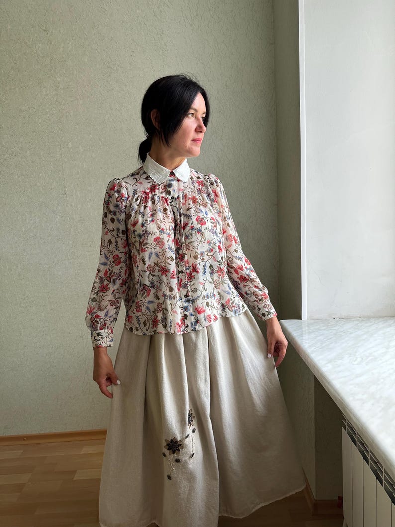 May include: A woman wearing a white collared blouse with a floral print and a beige skirt with embroidery. The blouse has long sleeves and a button-up front. The skirt is a-line and falls to the woman's ankles.