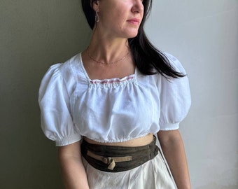 Pode incluir: Uma blusa branca de linho com mangas bufantes e um cinto de couro castanho. A blusa tem uma bainha bordada vermelha em volta do decote. A mulher está usando uma saia bege.