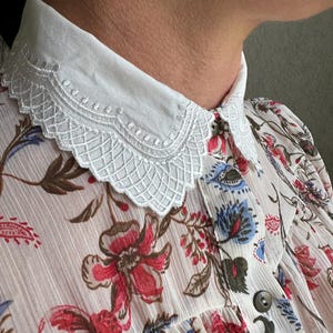 May include: A white cotton collar with a delicate lace trim. The collar is worn over a floral print blouse with a button closure.