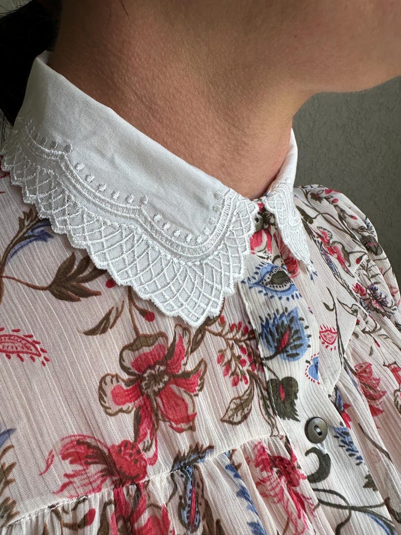 May include: A white cotton collar with a delicate lace trim. The collar is worn over a floral print blouse with a button closure.